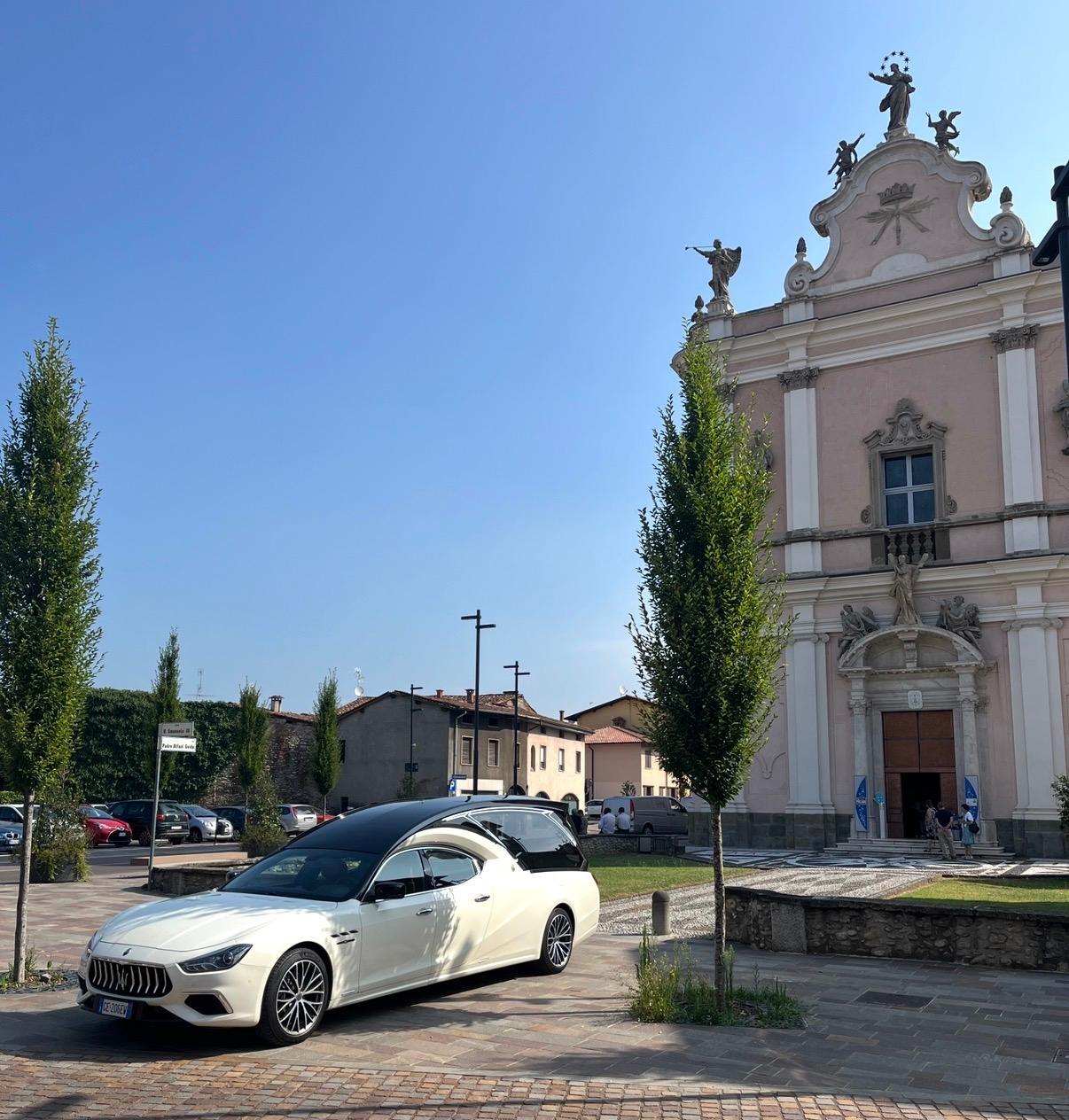 Carro funebre Maserati bianco dal tetto nero davanti a una chiesa barocca rosa con statue sulla facciata